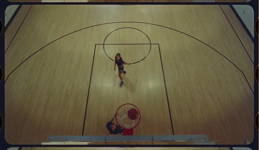 Slow motion, high-angle top-down shot of a man and woman playing one-on-one basketball, with the man successfully making a layup