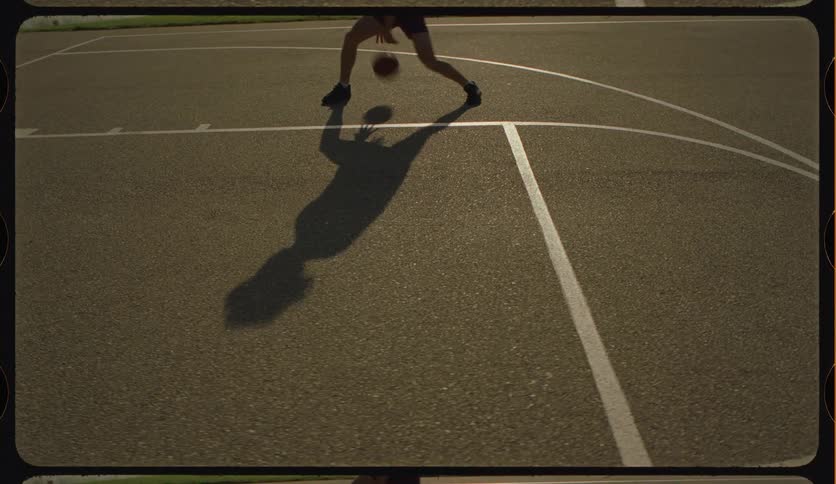 Slow-motion view of a woman's shadow as she dribbles a basketball on an outdoor court at sunset