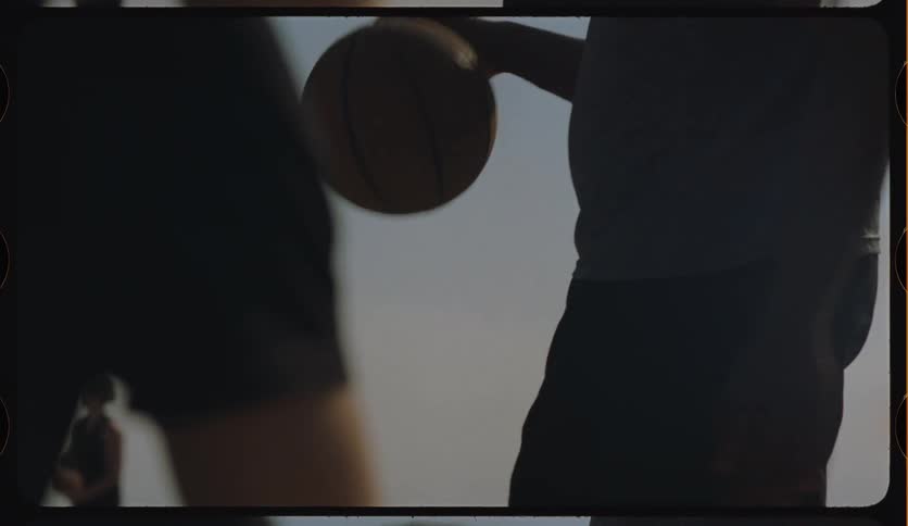A silhouetted basketball player dribbles against a defender in a slow-motion, cinematic shot with a retro film overlay