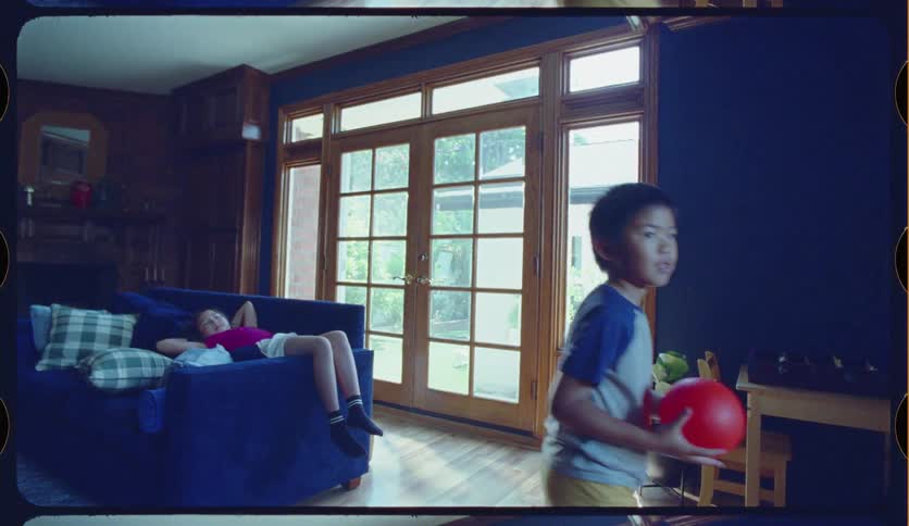 Slow motion shot of a young boy blowing up and playing catch with a red ball in a sunlit living room