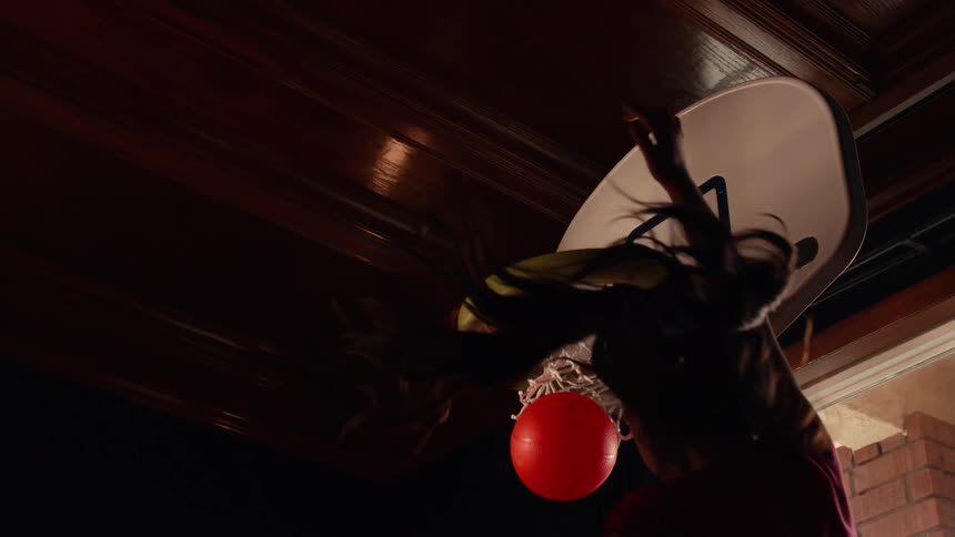 From a low angle, a silhouetted girl with long hair slam dunks a red ball into a toy basketball hoop in dramatic slow motion
