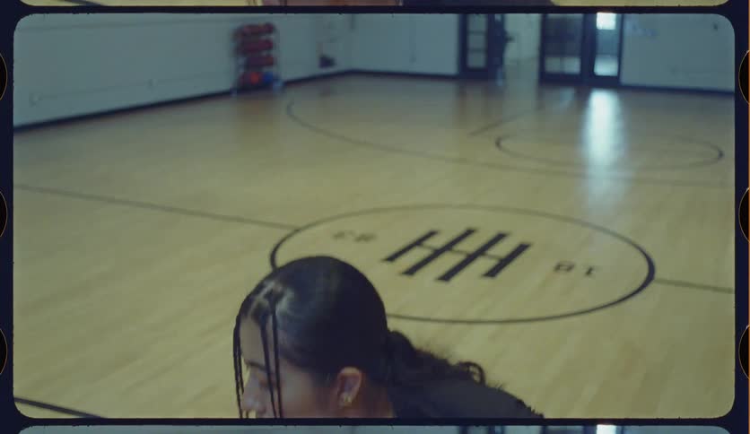 A focused female athlete with braided hair practices basketball drills on an indoor court with a vintage film aesthetic