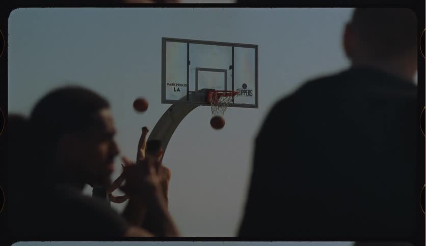 Silhouetted basketball players practice shooting hoops in slow motion on an outdoor court with a moody vintage film look