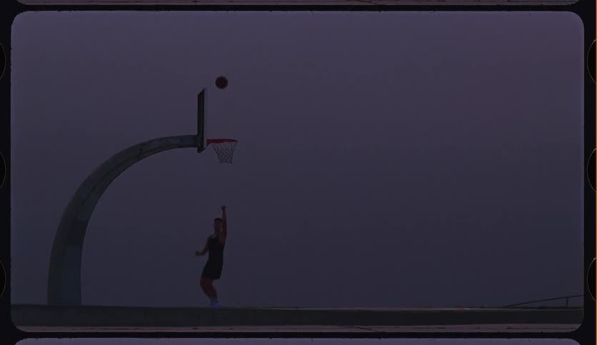 A man shoots and makes a basket in slow motion on a foggy outdoor court at dusk, framed with a vintage film look