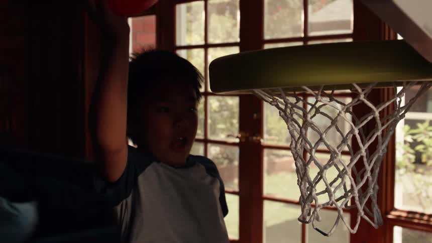 Young boy excitedly dunks a red ball into a toy basketball hoop in slow motion inside his home
