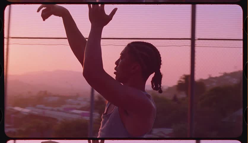 A young athletic man with braided hair shoots a basketball in slow motion on a picturesque outdoor court at sunset