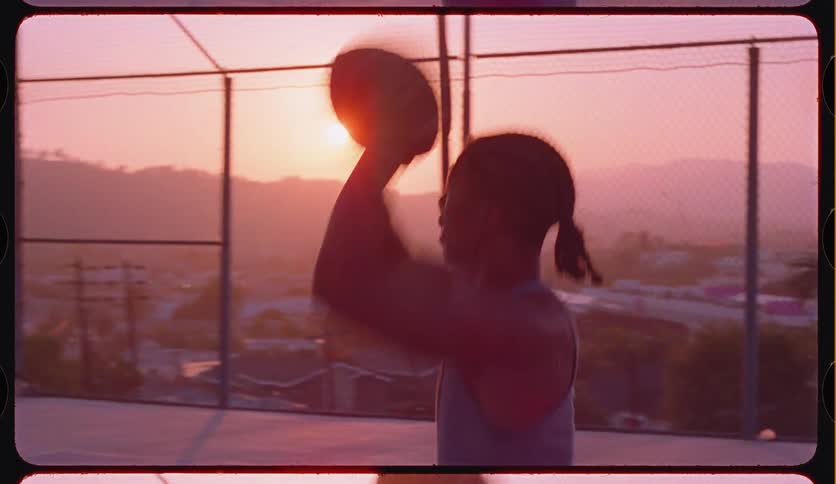 An athletic young man shoots a basketball in slow motion on an urban court against a vibrant sunset, captured with a vintage film aesthetic