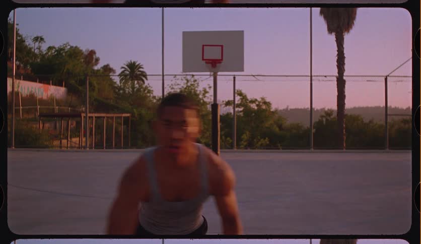 A determined man dribbles a basketball toward the camera on a vintage-looking outdoor court at sunset