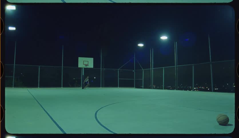 Young man practices basketball drills alone on an empty, illuminated outdoor court late at night, shot on 16mm film