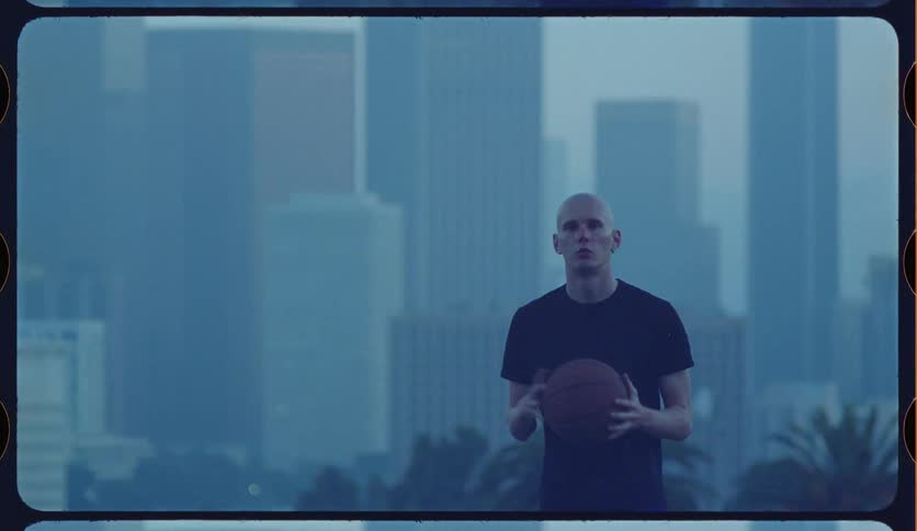 A serious basketball player stands with a ball, looking intently forward with a hazy blue city skyline in the background
