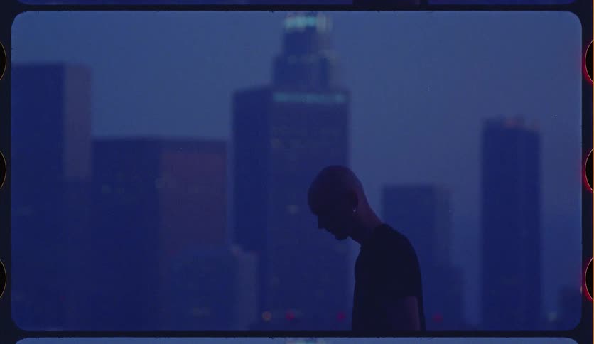 Moody cinematic shot of a man's silhouette holding a basketball against a hazy blue city skyline at dusk, captured on film