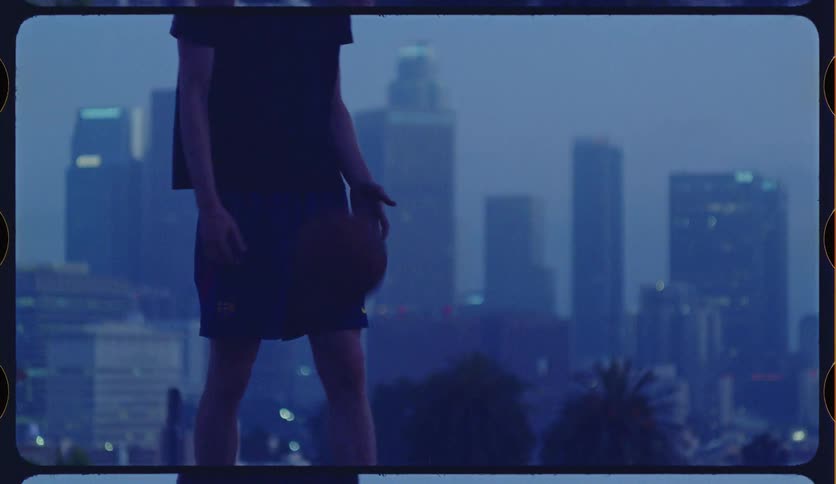A man practices his basketball dribbling skills in slow motion with the moody, blue-toned Los Angeles skyline visible in the background
