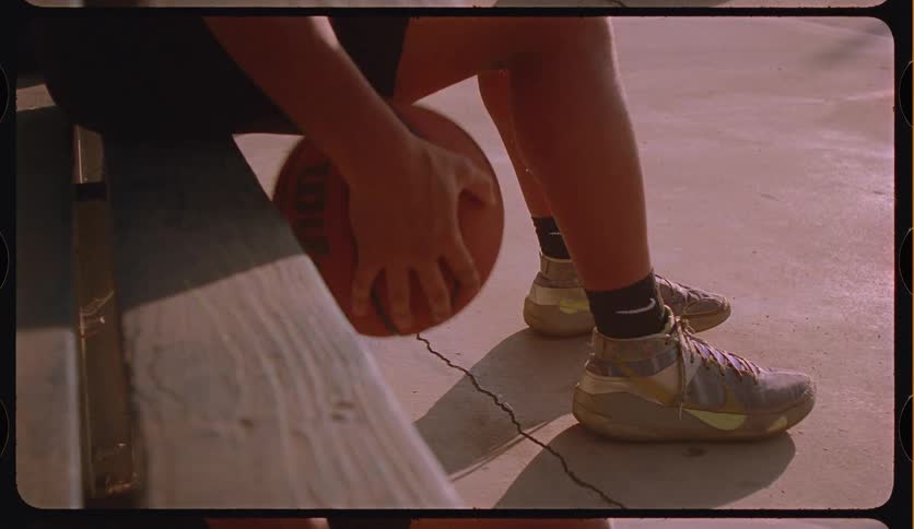 Vintage film-style shot of a player dribbling a basketball while sitting on a court-side bench in golden sunlight