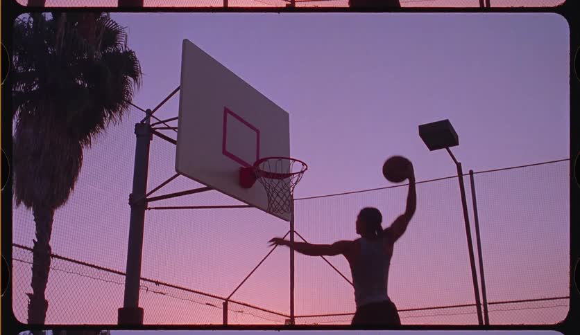 A silhouetted man dunks a basketball in slow motion on an outdoor court against a stunning purple and pink sunset
