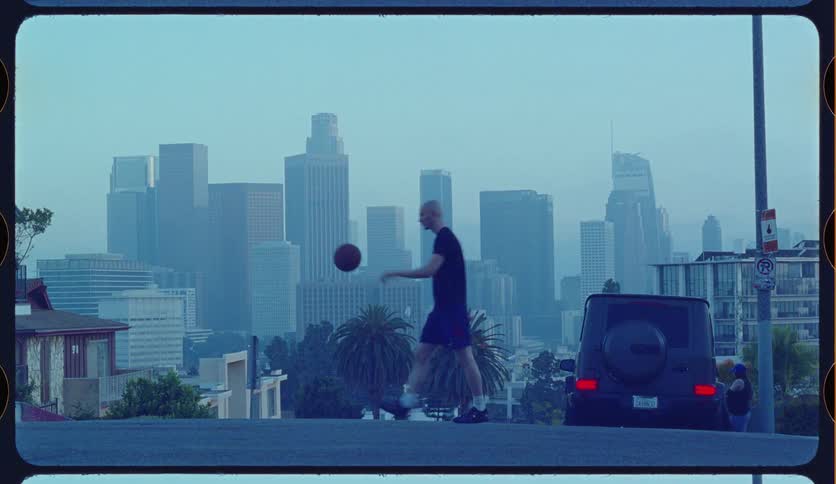 A man bounces a basketball in slow motion on a street with a scenic, hazy view of the downtown Los Angeles skyline
