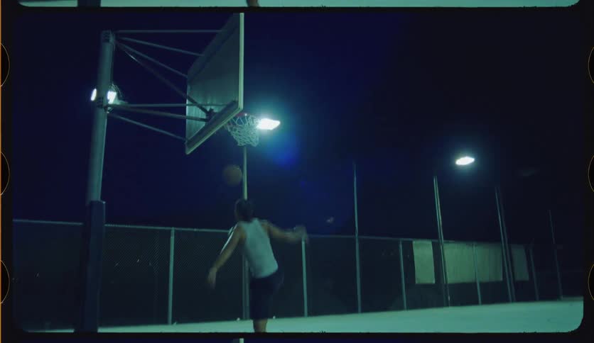 A basketball player slam dunks the ball in slow motion on an empty outdoor court at night, framed like a vintage film