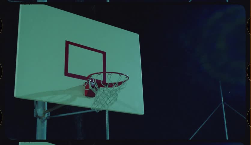 A basketball player dunks a ball in slow motion on an outdoor urban court at night with a vintage film look
