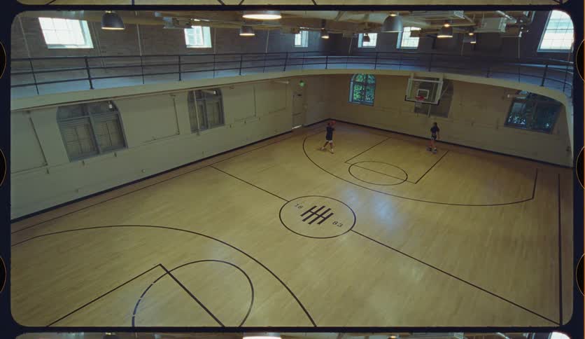 A high-angle wide shot shows two people casually playing basketball on a vintage indoor court with a retro film frame aesthetic