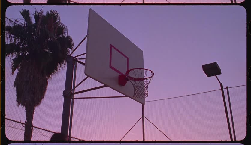 A basketball player slam dunks a ball in slow motion on an outdoor court, silhouetted against a beautiful pink and purple sunset sky