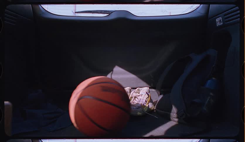 Basketball gear, including shoes and a ball, rolls around in the trunk of a moving car on a sunny day