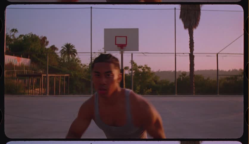 A young man dribbles a basketball towards the camera on an outdoor court at sunset with a vintage film look