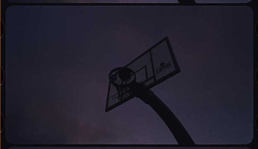 A low angle, slow motion shot of a person's silhouette scoring a basket against the purple twilight sky