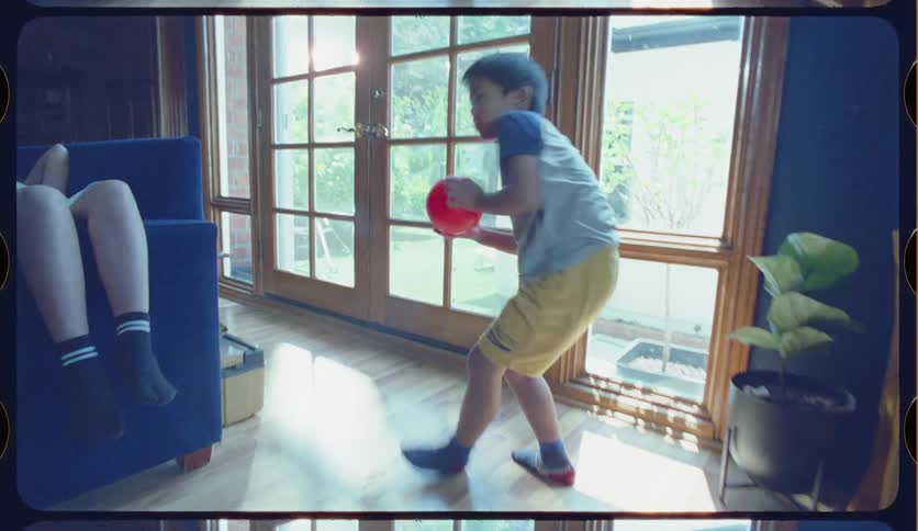 With a nostalgic film aesthetic, a young boy intently throws a red ball in his living room as his sister naps on the sofa