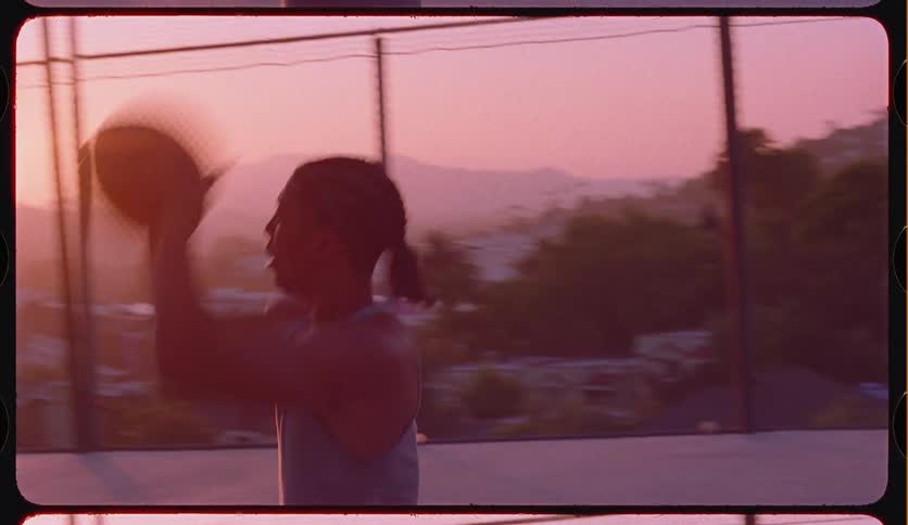 A young basketball player with braided hair shoots a hoop in slow motion on an outdoor court during a beautiful sunset