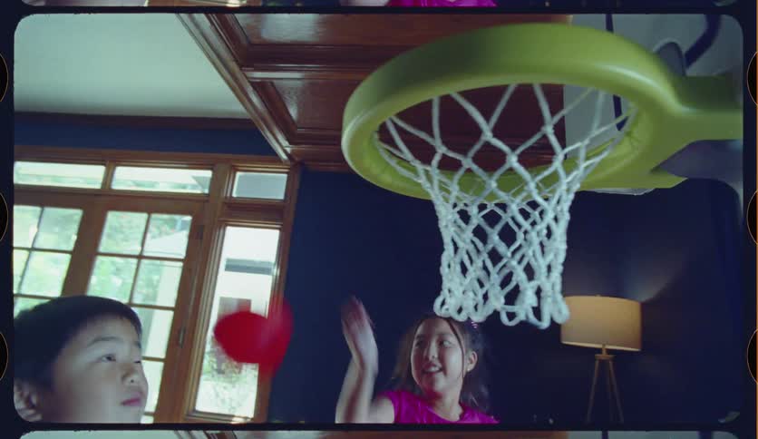 A young boy and girl joyfully play with a toy basketball hoop in their home, filmed in a nostalgic slow-motion style