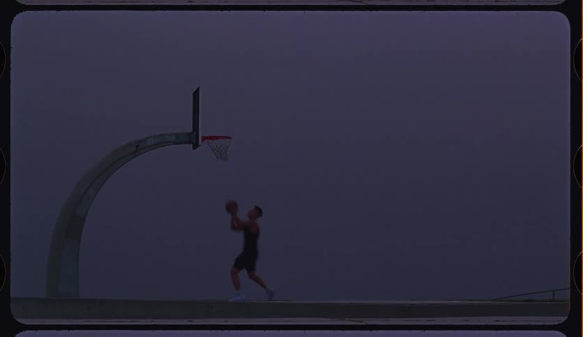 A basketball player makes a layup in slow motion on a moody outdoor court at dusk, shot on vintage film