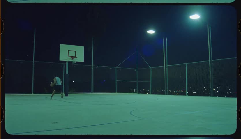 A solitary basketball player practices dribbling and scores a layup on an empty, illuminated court with city lights in the background at night