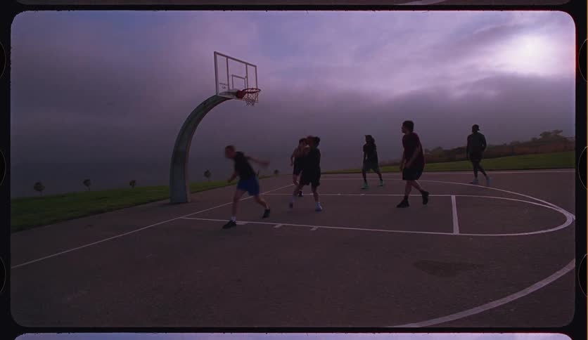 Slow motion footage of men playing basketball on an outdoor court with a dramatic cloudy sky