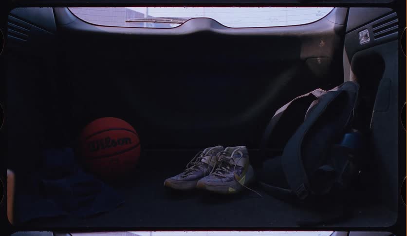 A basketball and a pair of worn-out sneakers sit in the back of a moving car, captured in a gritty, nostalgic film style
