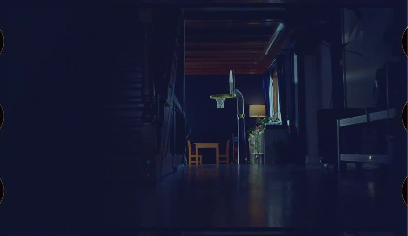 A young brother and sister take turns playing with a toy basketball and hoop in a dimly lit playroom at home