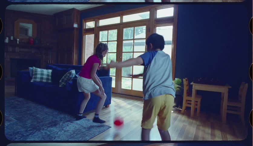 Energetic siblings laugh and play together, dribbling a red ball and shooting hoops in their playroom in this slow-motion, vintage-style home video