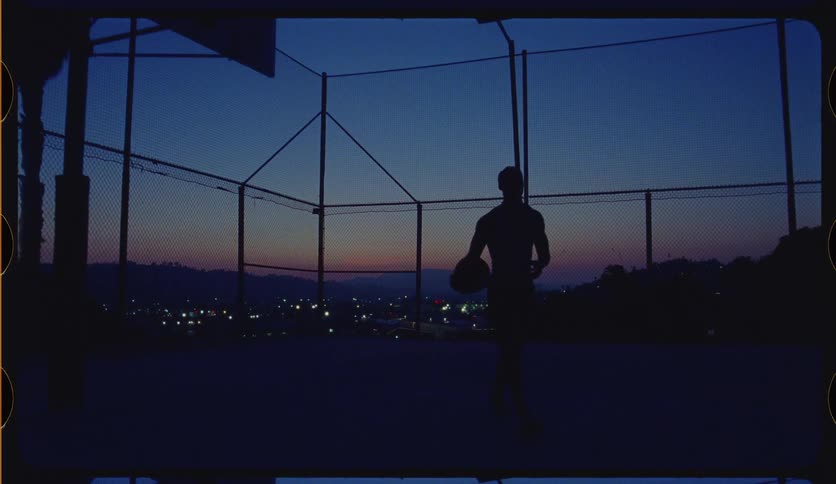 Slow motion silhouette of a person walking across an outdoor basketball court with a beautiful city view at dusk