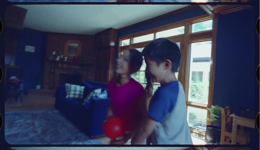 Vintage-style, slow-motion footage captures two young siblings laughing and taking turns shooting hoops on a toy basketball set in their living room