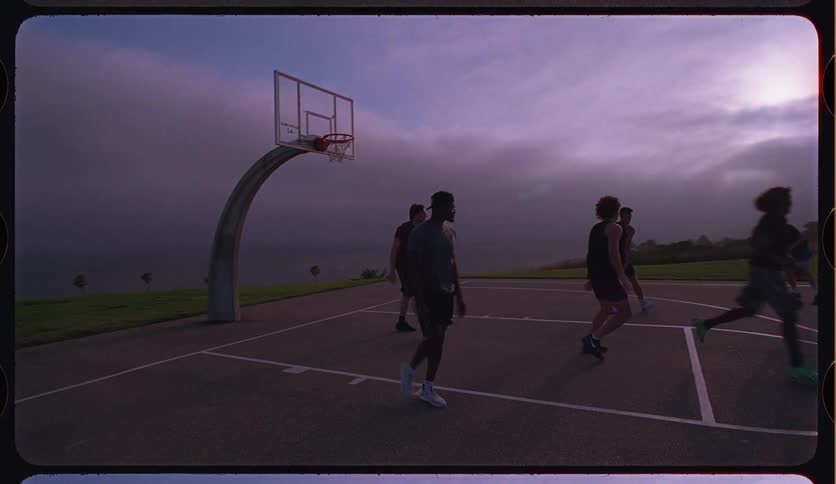 A group of friends play a game of basketball in slow motion on an outdoor court by the ocean during a cloudy sunrise