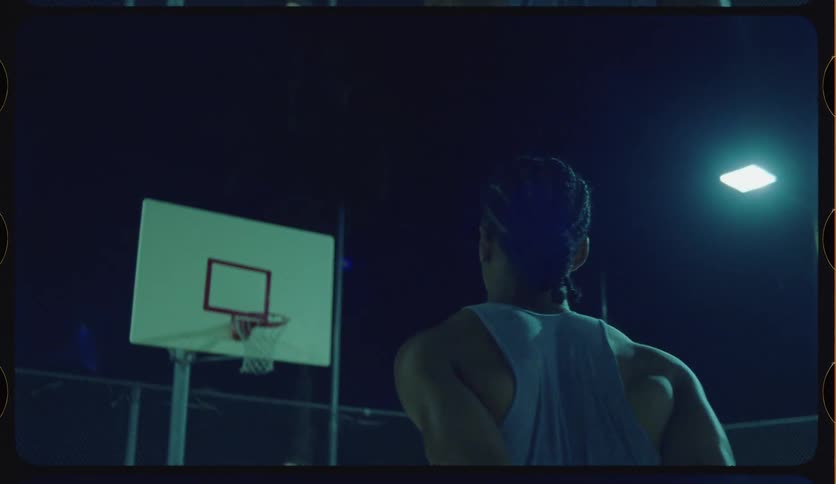A lone basketball player in a tank top makes a basket in slow motion on a dark outdoor court at night