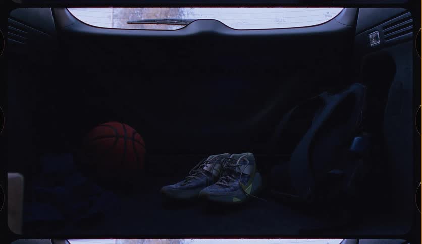 A basketball, sneakers, and sports bag sit in the back of a car as sunlight and shadows from passing trees wash over them