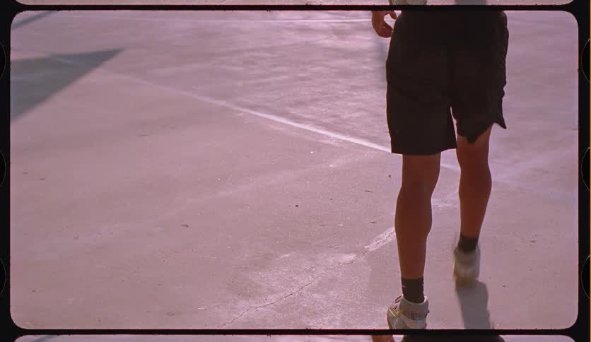 A focused young man with braided hair dribbles a basketball on a sunny court, with the shot following his shadow across the concrete