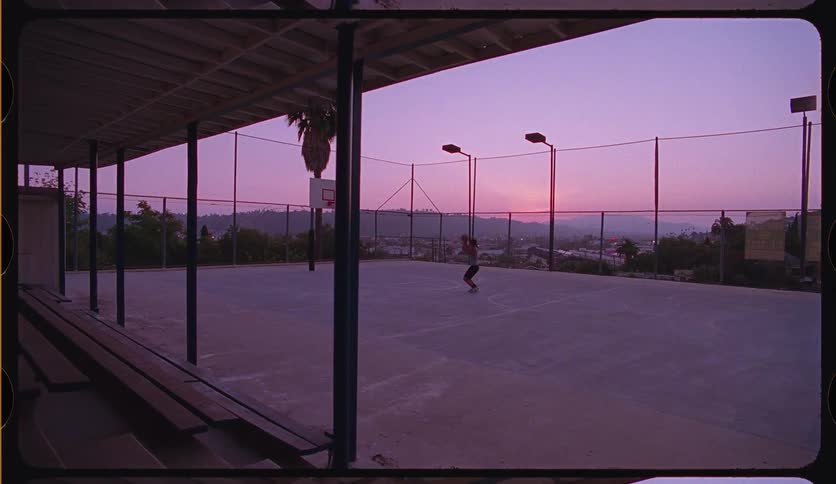 A young man practices basketball alone on an outdoor court against the beautiful backdrop of a purple and pink sunset sky