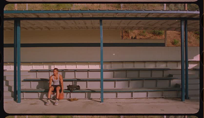 A contemplative young man sits alone on empty bleachers, slowly bouncing a basketball in a vintage-style shot