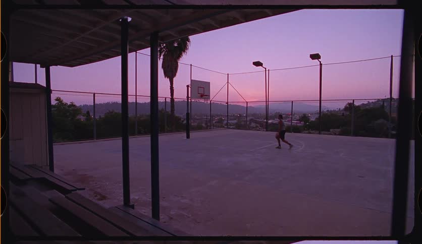 Slow dolly shot of a man playing basketball alone on an outdoor court against the backdrop of a vibrant purple sunset