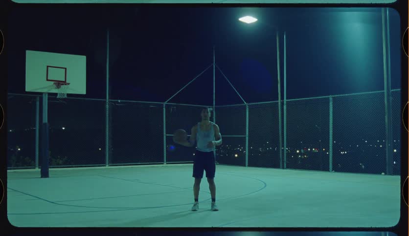 A basketball player stands alone and focused on an outdoor court at night, illuminated by a single light with the city skyline in the background