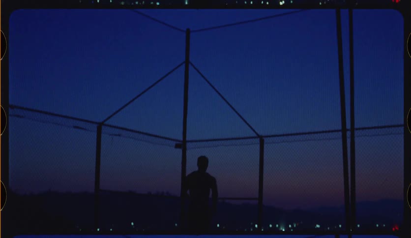Vintage-style footage of a lone basketball player's silhouette practicing on a fenced court as day turns to night over the city