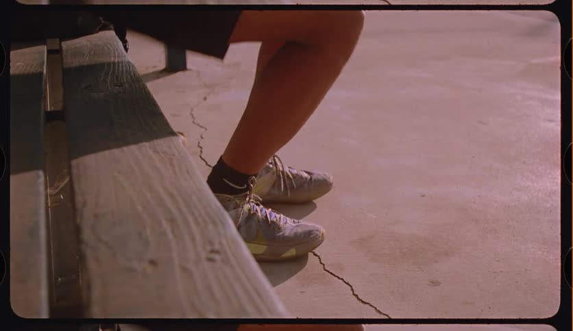 Vintage-style footage of a basketball player tying his shoes on a bench and then walking away, dribbling the ball on a sunny court