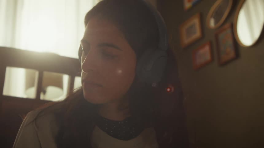 A young woman with headphones on listens to music with her eyes closed, relaxing in a sunlit room