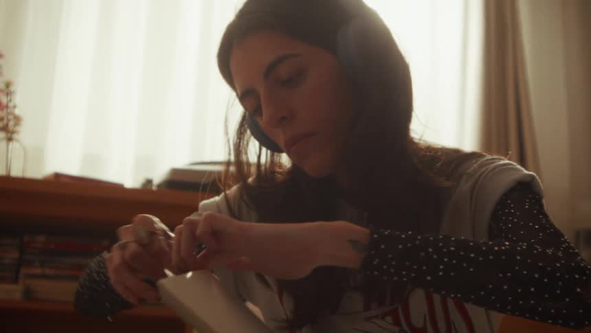 Contemplative young woman in headphones takes a vinyl record out of its sleeve in slow motion with a warm sun flare in her room