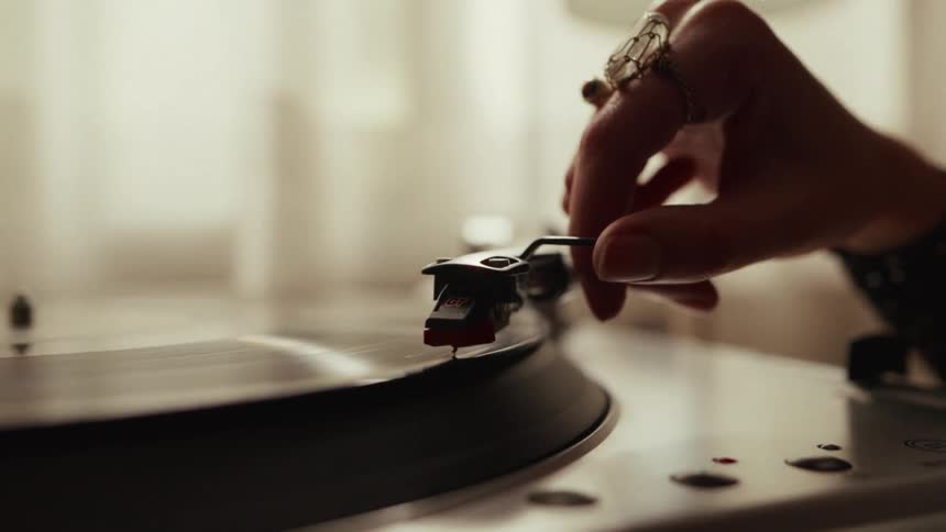 A person's hand with rings gently lowers the stylus onto a spinning vinyl record on a turntable